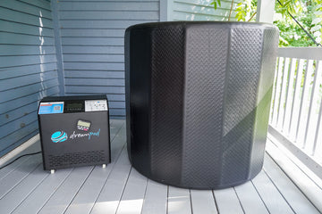 The upright Dreampod Cold Plunge Barrel commercial cylindrical tub standing alongside its external water chiller on a wooden deck with a blue wall and white railing in the background.